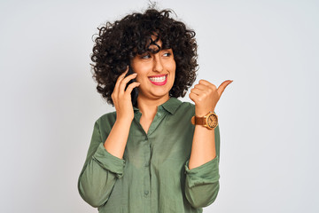 Young arab woman with curly hair talking on smartphone over isolated white background pointing and showing with thumb up to the side with happy face smiling