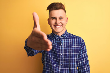 Young handsome man wearing casual shirt standing over isolated yellow background smiling friendly offering handshake as greeting and welcoming. Successful business.