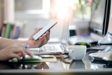 Croopped shot of man hand using smartphone while sitting at office desk and working with laptop computer.