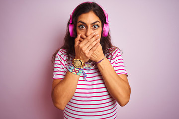 Young beautiful woman listening to music using headphones over isolated pink background shocked covering mouth with hands for mistake. Secret concept.