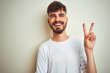 Fototapeta premium Young man with tattoo wearing t-shirt standing over isolated white background smiling with happy face winking at the camera doing victory sign. Number two.