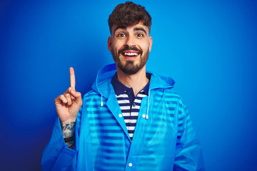 Young handsome man wearing rain coat standing over isolated blue background pointing finger up with successful idea. Exited and happy. Number one.
