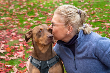 Blond woman kissing Doberman mix dog, outside on green lawn covered in red fall leaves