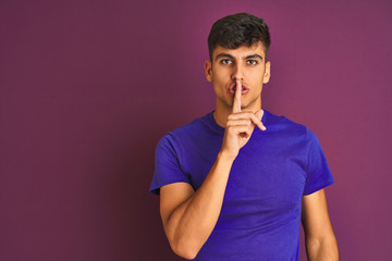 Young indian man wearing t-shirt standing over isolated purple background asking to be quiet with finger on lips. Silence and secret concept.