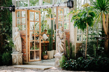 In the corner of the picture, you can see a glass house inside the house. You can see a cactus in a pot with many species of plants and many different kinds of plants.