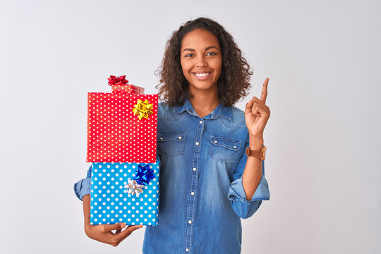 Young brazilian woman holding birthday gifts standing over isolated white background surprised with an idea or question pointing finger with happy face, number one