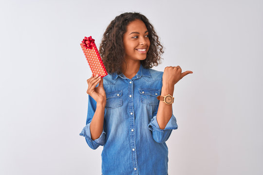 Young brazilian woman holding valentine gift standing over isolated white background pointing and showing with thumb up to the side with happy face smiling