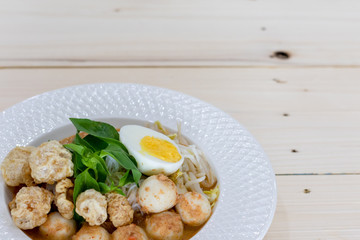 Asian Food Rice Noodles in Fish Curry With boiled egg, fish balls, pork snack, basil, bean sprouts on top, placed on a wooden table.