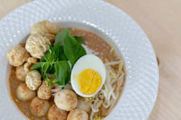 Thai Food - Rice Noodles in Fish Curry With boiled egg, fish balls, pork snack, basil, bean sprouts on top, placed on a wooden table.