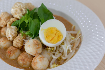 Thai Food Rice Noodles in Fish Curry With boiled egg, fish balls, pork snack, basil, bean sprouts on top, placed on a wooden table.