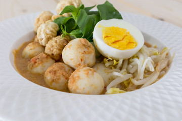 Thai Food with Rice Noodles in Fish Curry With boiled egg, fish balls, pork snack, basil, bean sprouts on top, placed on a wooden table.