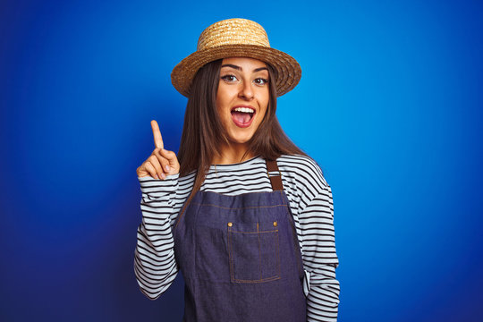Young beautiful baker woman wearing apron and hat standing over isolated blue background pointing finger up with successful idea. Exited and happy. Number one.