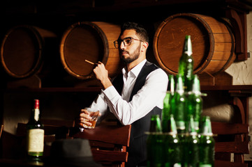 Handsome well-dressed arabian man with glass of whiskey and cigar posed at pub.