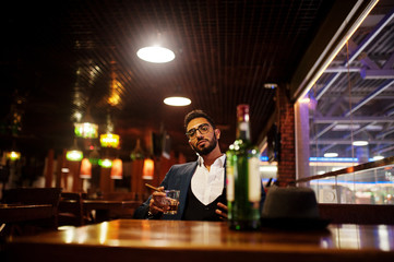 Handsome well-dressed arabian man with glass of whiskey and cigar hold mobile phone,  posed at pub.