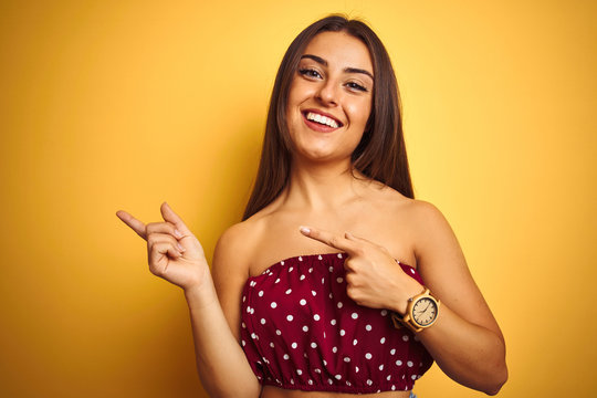 Young beautiful woman wearing red t-shirt standing over isolated yellow background smiling and looking at the camera pointing with two hands and fingers to the side.