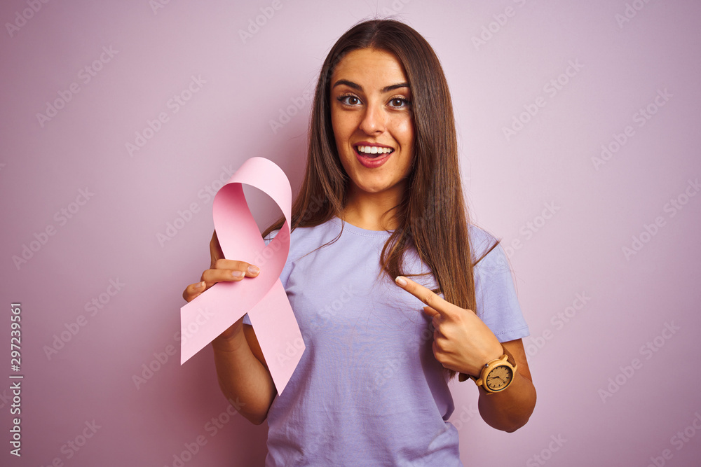 Wall mural Young beautiful woman holding cancer ribbon standing over isolated pink background very happy pointing with hand and finger