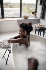 Beaming curly woman feeling cheerful while lying in foamy bath