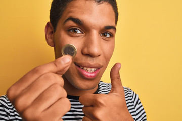 Young handsome arab man holding euro coin standing over isolated yellow background happy with big smile doing ok sign, thumb up with fingers, excellent sign