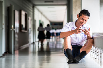 Fototapeta na wymiar Asian male high school student in a white uniform, who is a lot of addicted to games. He is playing exciting games on their mobile phones and sitting in the school.