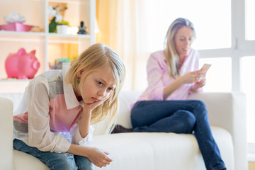 Mother is texting while her little girl  unhappy sitting next to her on couch