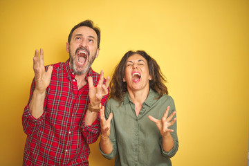 Beautiful middle age couple over isolated yellow background crazy and mad shouting and yelling with aggressive expression and arms raised. Frustration concept.