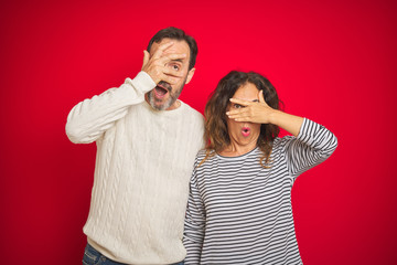 Fototapeta premium Beautiful middle age couple wearing winter sweater over isolated red background peeking in shock covering face and eyes with hand, looking through fingers with embarrassed expression.