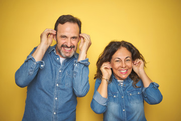 Beautiful middle age couple together standing over isolated yellow background Smiling pulling ears with fingers, funny gesture. Audition problem