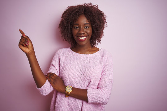 Young african afro woman wearing sweater standing over isolated pink background with a big smile on face, pointing with hand and finger to the side looking at the camera.