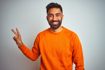 Fototapeta premium Young indian man wearing orange sweater over isolated white background smiling with happy face winking at the camera doing victory sign. Number two.