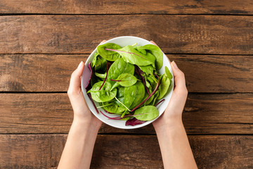 Overhead shot of woman’s hands holding green salad in bowl on rustic wooden background. Close up