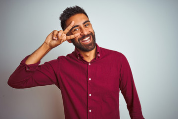 Fototapeta premium Young indian man wearing red elegant shirt standing over isolated grey background Doing peace symbol with fingers over face, smiling cheerful showing victory