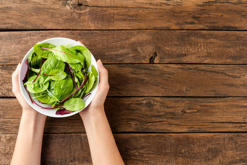 Overhead shot of female hands holding green salad in bowl on rustic wooden background with copyspace