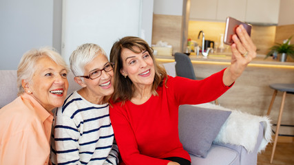 Obraz premium Beautiful senior women having fun taking selfies at home. Senior friends using a Phone. Senior women having fun with smartphone making selfies. Capturing their friendship