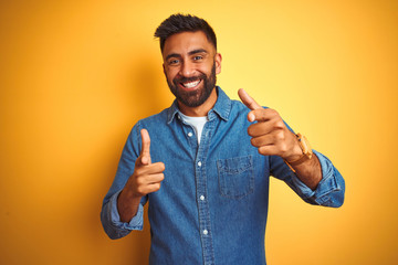 Young indian man wearing denim shirt standing over isolated yellow background pointing fingers to...