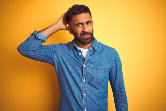 Young indian man wearing denim shirt standing over isolated yellow background confuse and wonder about question. Uncertain with doubt, thinking with hand on head. Pensive concept.