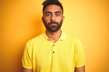 Obraz premium Young indian man wearing polo standing over isolated yellow background with serious expression on face. Simple and natural looking at the camera.