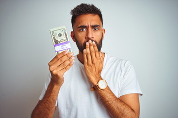 Young indian man holding dollars standing over isolated white background cover mouth with hand shocked with shame for mistake, expression of fear, scared in silence, secret concept