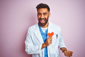 Young indian doctor man holding paper heart standing over isolated pink background screaming proud...