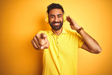 Arab indian hispanic man listening to music using headphones over isolated yellow background pointing with finger to the camera and to you, hand sign, positive and confident gesture from the front
