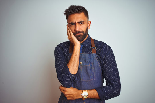 Young Indian Employee Man Wearing Apron Uniform Standing Over Isolated White Background Thinking Looking Tired And Bored With Depression Problems With Crossed Arms.