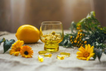 Traditional medicine background with dry chamomile flowers in glass cup, blurred lemon at burlap sack with omega 3 capsules, tansy, calendula at the table