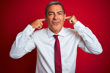 Handsome middle age businessman standing over isolated red background covering ears with fingers with annoyed expression for the noise of loud music. Deaf concept.