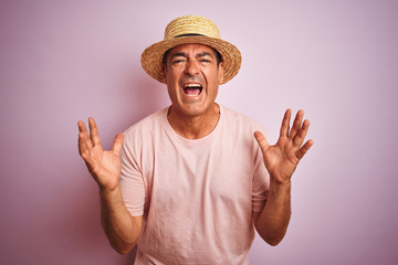 Handsome middle age man wearing summer hat standing over isolated pink background crazy and mad shouting and yelling with aggressive expression and arms raised. Frustration concept.