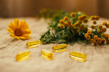 Obraz na płótnie Canvas Close up fish oil capsules with calendula flower and tansy at background on linen fabric at the table, copy space, soft focus
