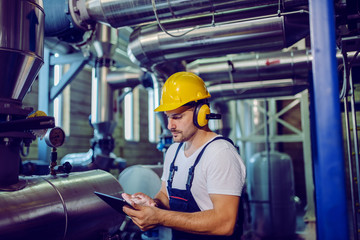 Focused plant worker in overalls, with protective helmet on head and antiphons on ears using tablet...