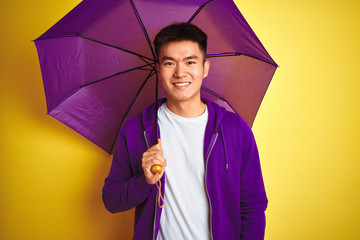 Asian chinese man wearing purple umbrella standing over isolated yellow background with a happy face standing and smiling with a confident smile showing teeth