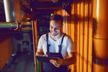 Smiling hardworking plant worker in coveralls standing and using tablet at night. Night shift.