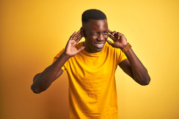 Fototapeta na wymiar Young african american man wearing casual t-shirt standing over isolated yellow background Trying to hear both hands on ear gesture, curious for gossip. Hearing problem, deaf