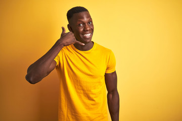 Young african american man wearing casual t-shirt standing over isolated yellow background smiling doing phone gesture with hand and fingers like talking on the telephone. Communicating concepts.