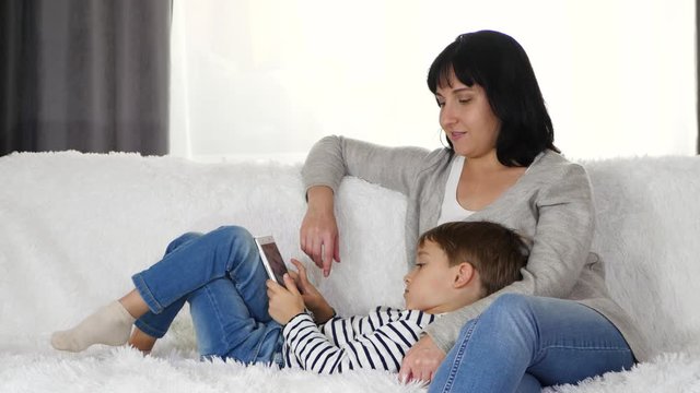 A happy mother and a young child spend time with a tablet while sitting on the couch at home. Mother hugging her son. The concept of a happy modern family.